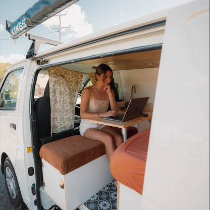 Woman using a laptop inside a converted solar-powered campervan on a wooden desk.